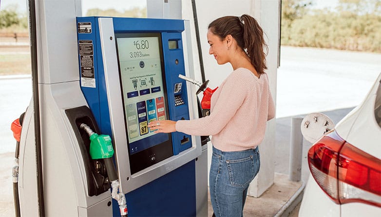 A woman at a Wayne gas pump, specifically the Anthem UX fuel dispenser, about to pump fuel using the interactive display.